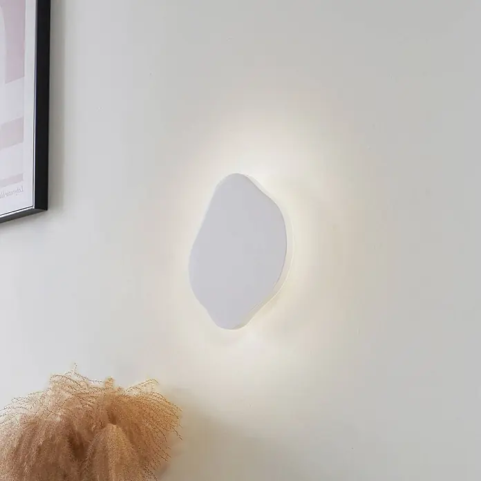 Lindby LED Wandleuchte innen in weiß, up down Wandlampe -  - Weiße, organisch geformte Wandleuchte mit warmem Licht, montiert an einer hellen Wand neben einem gerahmten Bild und Trockengras.