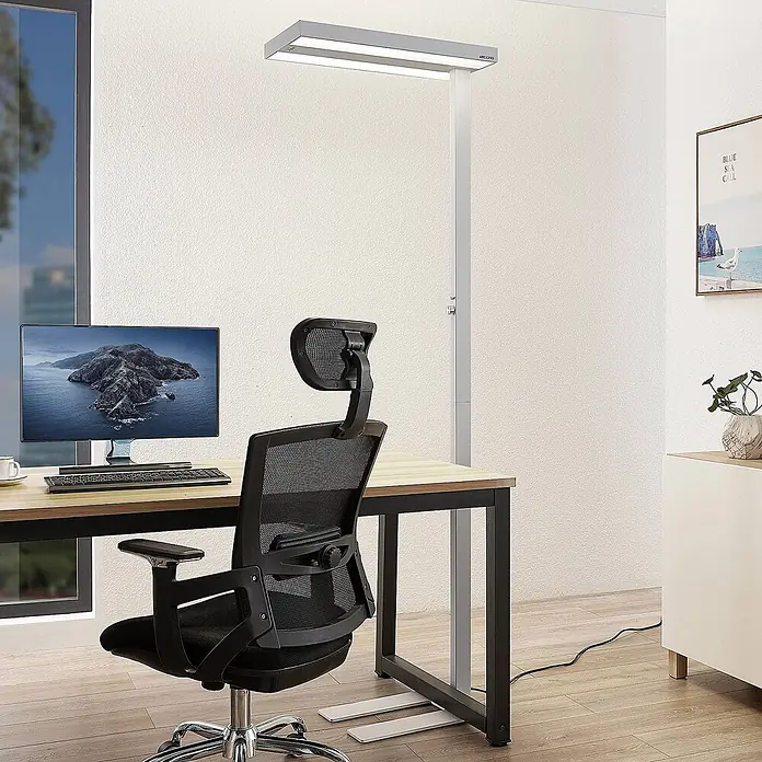 Arcchio LED Büro Stehlampe dimmbar, Bewegungsmelder, Tageslichtsensor -  - Moderne silberne Stehleuchte mit rechteckigem Kopf, die einen Büroschreibtisch mit Computer und Stuhl beleuchtet.