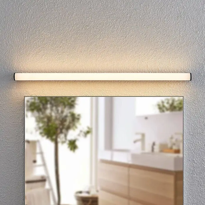 Lindby LED Wandleuchte innen in schwarz matt, weiß, Wandlampe -  - Lange, lineare LED-Leuchte mit schwarzen Enden, die warmweißes Licht abgibt, horizontal über einem verschwommenen Badezimmerspiegel montiert.
