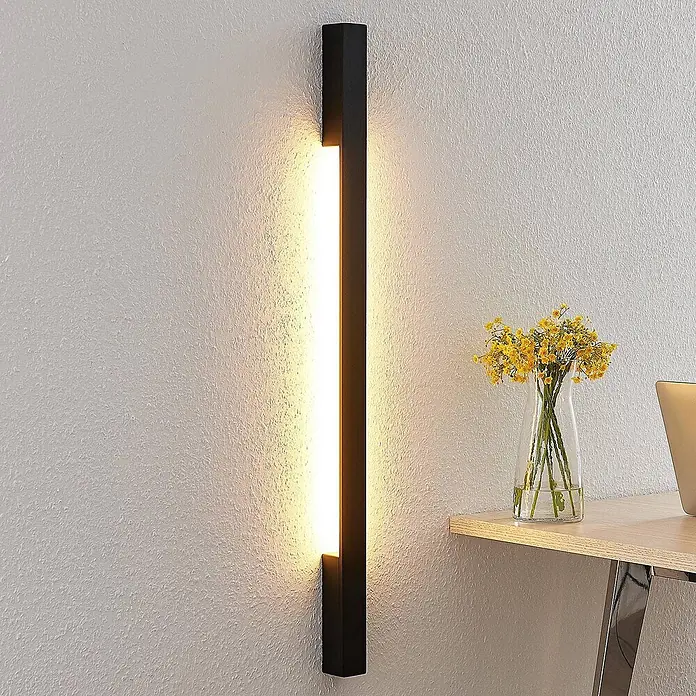 Arcchio LED Wandleuchte innen in schwarz, Wandlampe -  - Vertikale schwarze LED-Wandleuchte beleuchtet eine strukturierte Wand über einem Schreibtisch mit gelben Blumen.