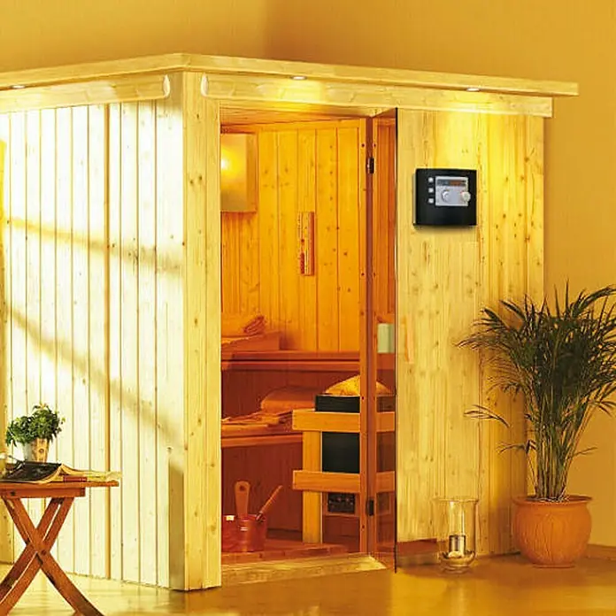 Karibu Elementsauna Rodin - Mit 9 kW Ofen mit integrierter Steuerung, Ohne Dachkranz, L x B x H: 196 x 196 x 192 cm - Eine gemütliche Holzsauna mit Steuerungspanel an der Wand und Entspannungsecke im Vordergrund.