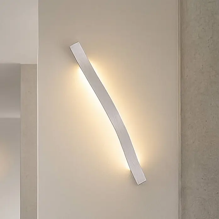 Lindby Wandleuchte Alvon - alu / grau / zink - Eine geschwungene, silberne LED-Wandleuchte aus gebürstetem Metall, die warmes indirektes Licht abgibt.