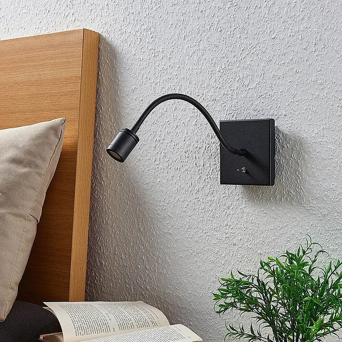 Lindby LED Wandleuchte innen in schwarz, Wandlampe -  - Schwarze Wandleseleuchte mit flexiblem Schwanenhals, neben einem Holzbett mit Buch und Pflanze montiert.