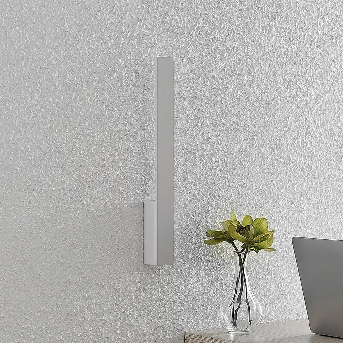 Arcchio LED Wandleuchte innen in weiß, Wandlampe -  - Weiße rechteckige Wandleuchte an einer strukturierten Wand, neben einer Vase mit grünen Blumen und einem Laptop auf einem Schreibtisch.