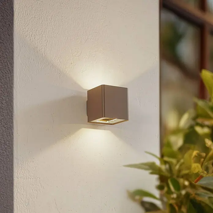 Lucande LED Wandleuchte aussen in braun, Aussenlampe Wand -  - Eine quadratische, dunkelgraue Außenwandleuchte an einer strukturierten Wand, die warmes Licht nach oben und unten abstrahlt.
