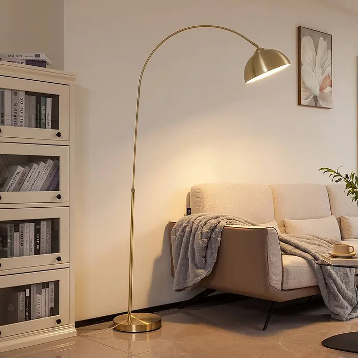 Lindby Stehlampe in messing matt, Bogenlampe Wohnzimmer -  - Goldene Bogen-Stehleuchte mit Kuppelschirm, die warmes Licht spendet, neben einem Sofa im Wohnzimmer.