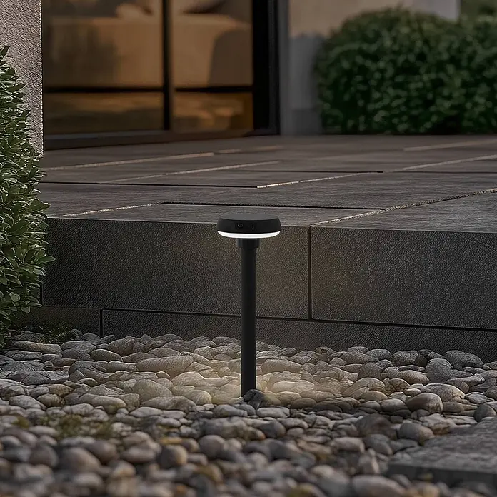 Lindby Erdspießleuchte Brinca -  - Eine schwarze Wegeleuchte mit weißem Licht beleuchtet Kieselsteine neben einer modernen Terrasse.