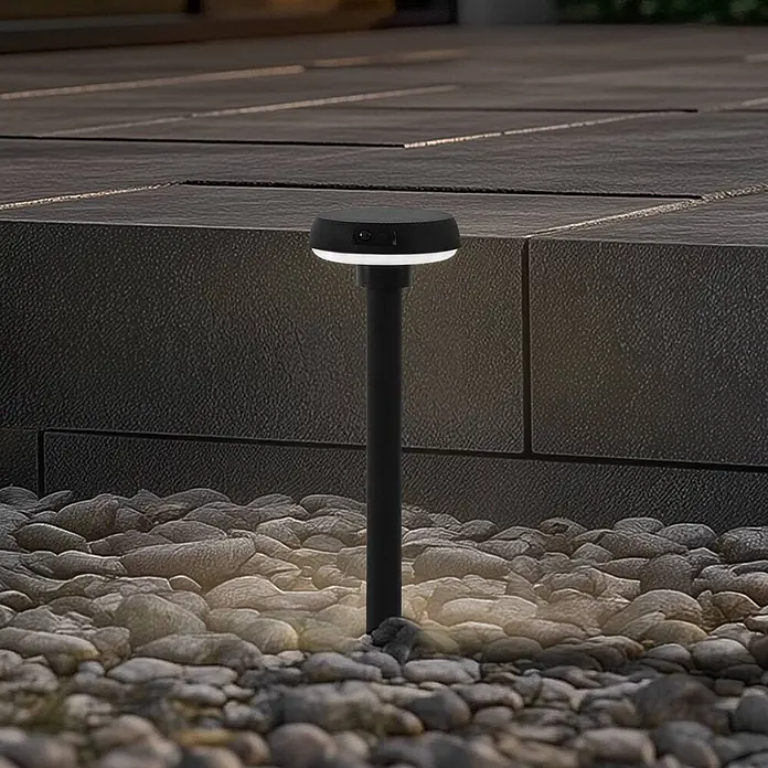 Lindby Erdspießleuchte Brinca -  - Schwarze Solar-Gartenwegleuchte mit leuchtendem weißem Ring, wirft warmes Licht auf Zierkiesel.