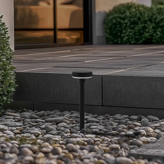 Lindby Erdspießleuchte Brinca -  - Schwarze Gartenwegleuchte mit rundem Kopf und weißem Ring, steht zwischen grauen Kieseln neben einem gepflasterten Bereich.