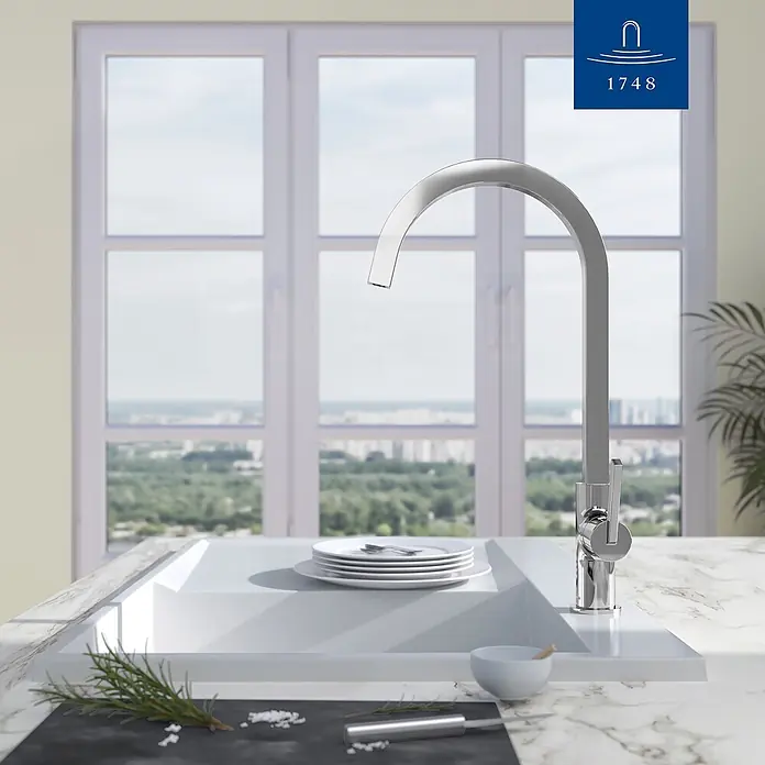 Villeroy & Boch Spültischarmatur - Chrom, Hochglänzend - Chrom-Küchenarmatur über modernem weißen Spülbecken mit Geschirr, vor hellem Fenster mit Stadtblick.