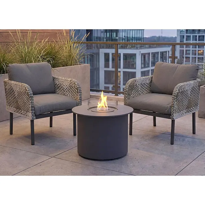 Mendler Loungemöbel-Set HWC-P80 - 3 -tlg., Rattan, Dunkelgrau, Dunkelgrau - Zwei graue Outdoor-Sessel mit Geflecht und ein runder grauer Feuertisch mit Glasflamme auf einem modernen Balkon.