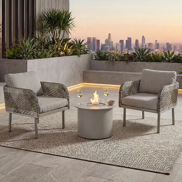 Mendler Loungemöbel-Set HWC-P80 - 3 -tlg., Rattan, Creme-beige, Creme-beige - Zwei graue Outdoor-Sessel mit geflochtenen Seiten und ein runder Feuertisch auf einem Teppich, auf einem Balkon mit Stadtblick.