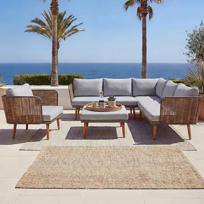Mendler Loungemöbel-Set HWC-L31 - 4 -tlg., Textil, Hellgrau, Hellgrau - Outdoor-Sofa und Sessel mit hellgrauen Kissen und braunen Geflechtrahmen auf einer Terrasse mit Meerblick, mit zwei Naturfaserteppichen.
