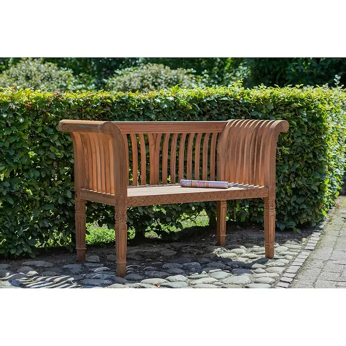 Bene Living Gartenbank Newport - Teakholz, Natur - Eine hellbraune Gartenbank aus Holz mit geschnitzten Beinen und Lattenrückenlehne auf einem Kopfsteinpflasterweg neben einer grünen Hecke.