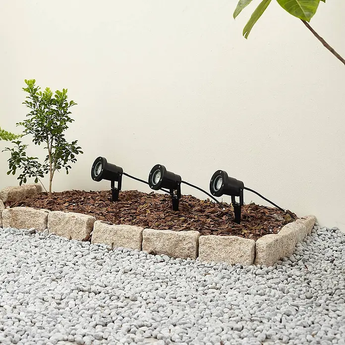 Lindby Gartenstrahler mit Erdspieß in schwarz, Strahler Garten -  - Drei schwarze Gartenstrahler mit Kabeln beleuchten ein Gartenbeet mit Rindenmulch, umrandet von Stein und Kies.