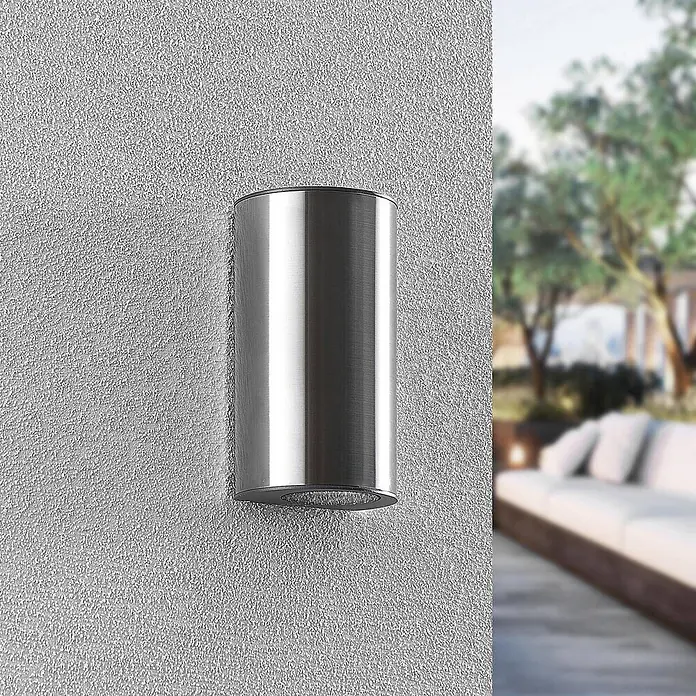 Prios Außenwandleuchte Zavallina - alu / grau / zink, IP54 - Zylindrische Wandleuchte aus gebürstetem Silber für den Außenbereich, montiert an einer strukturierten Wand mit verschwommener Terrasse im Hintergrund.