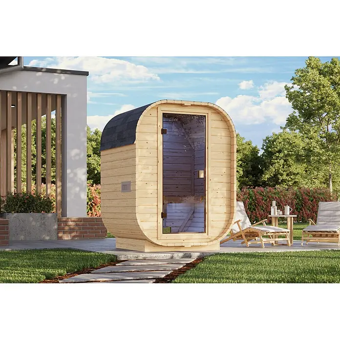 Karibu Saunahaus Sam - Mit Ofen, L x B x H: 140 x 158 x 205 cm - Helle Holz-Fasssauna mit dunklem Dach und Glastür, auf einer Terrasse neben einem Haus mit Liegestühlen.