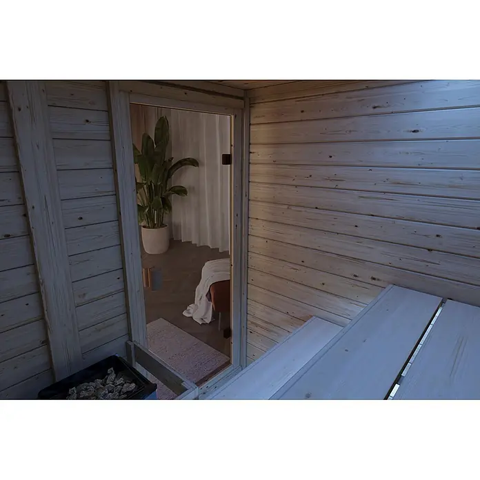 Karibu Elementsauna Oda - Mit Ofen, L x B x H: 145 x 148 x 190,5 cm - Heller Holzsauna-Innenraum mit Glastür und Blick in einen Raum mit Pflanze und Bett.