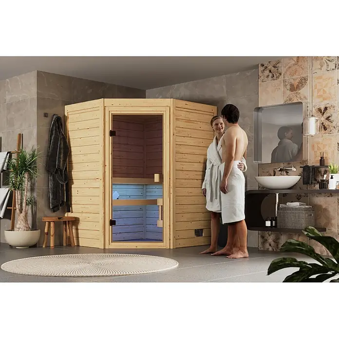 Karibu Elementsauna Oda Eck - Mit Ofen, L x B x H: 145 x 148 x 190,5 cm - Helle Ecksauna aus Holz mit Glastür in einem modernen Bad, daneben ein Paar in Bademänteln.