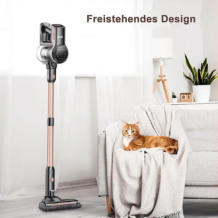 INSE Akku-Staubsauger - 1 Akku, 2,5 Ah, Behältervolumen: 2 l - Schwarz-roségoldener Stielstaubsauger steht aufrecht in einem Wohnzimmer neben einem Sofa mit Katze, was sein freistehendes Design hervorhebt.
