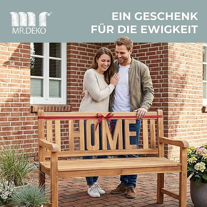 Bene Living Gartenbank Swindon - Teakholz, Natur - Holzgartenbank mit 'HOME'-Schriftzug in der Rückenlehne, mit roter Schleife verziert, auf einer Terrasse mit lächelndem Paar.