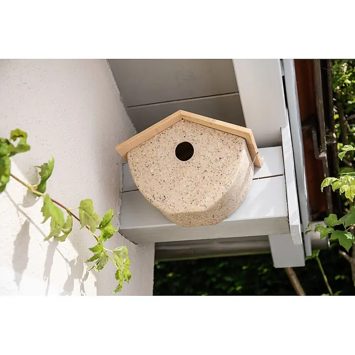 Windhager Nistkasten - 16 x 25 x 19,6 cm - Gesprenkeltes beiges Vogelhaus mit Holzdach, draußen an einem weißen Balken unter einem Überstand montiert.