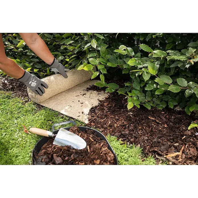 Windhager Unkrautmatte - L x B: 4 x 0,5 m - Hände in Gartenhandschuhen rollen eine Unkrautvliesmatte aus Naturfasern neben einer Hecke aus, mit Mulch und einer Schaufel.