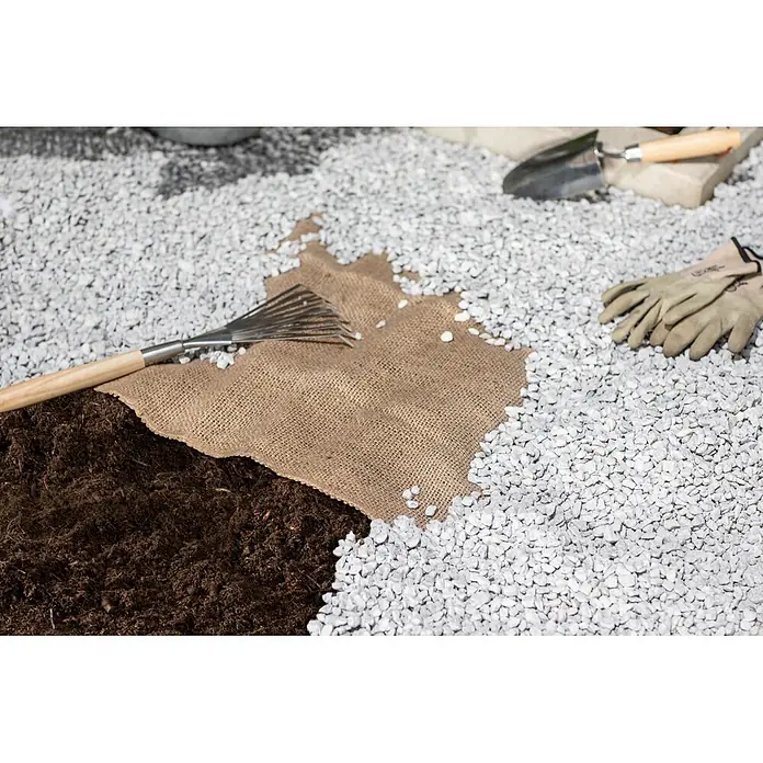Windhager Unterbodengewebe - Jute, L x H: 6 x 0,75 m, Unkrautschutz - Gartenarbeit mit Jute-Unkrautvlies, teilweise mit weißem Kies bedeckt, dazu Rechen, Schaufel und Handschuhe.