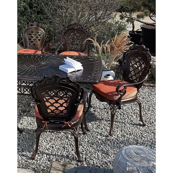 Beliani Gartenstuhl-Set LIZZANO - 2 Stk., Aluminium, Braun, B x T x H: 69 x 58 x 97 cm - Ein verziertes dunkelbronzefarbenes Terrassen-Esstisch-Set mit orangefarbenen Kissen auf einem Kiesweg, mit einem Buch auf dem Tisch.
