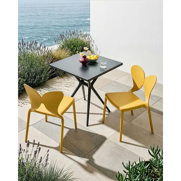 2er-Set Gartenstuhl LARISSA in Gelb -  - Zwei gelbe Outdoor-Stühle mit charakteristischer Rückenlehne und ein dunkelgrauer quadratischer Tisch auf einer Terrasse mit Meerblick, bereit für eine Mahlzeit.