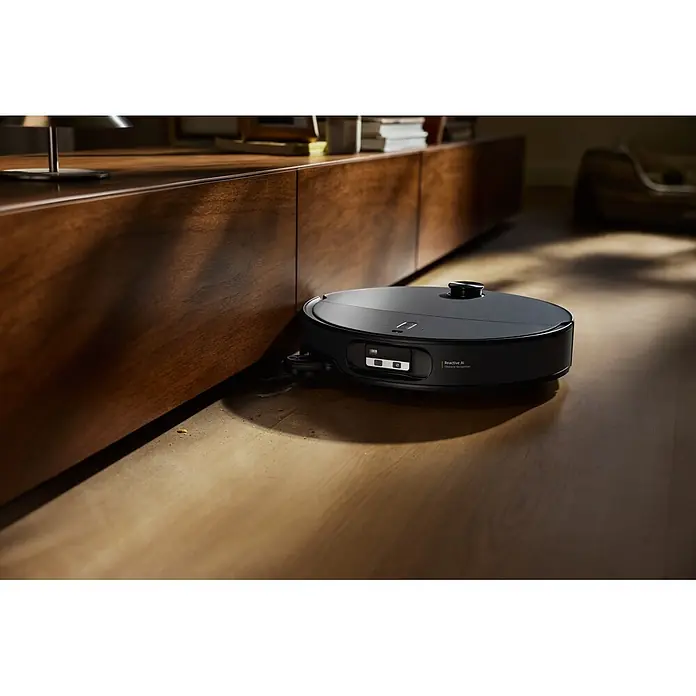 Roborock Saugwischroboter Qrevo Edge 2X - Saugkraft: 25.000 Pa, Akkulaufzeit: 188 min, Fassungsvermögen Roboter: 240 ml, Schwarz - Schwarzer Saugroboter reinigt einen Holzboden neben einem Schrank, mit sichtbarem Schmutz um seine Bürste.
