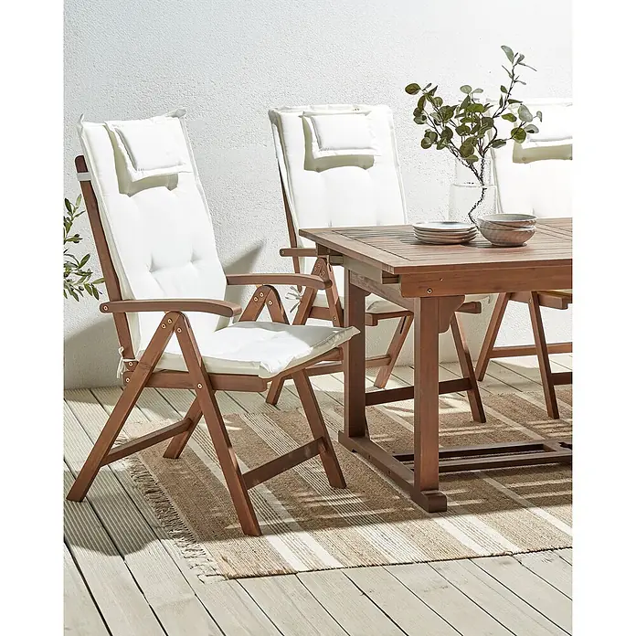 6er-Set Gartenstuhl AMANTEA in Braun/Weiß -  - Eine hölzerne Outdoor-Essgruppe mit vier Stühlen und einem Tisch, weißen Kissen und einer Dekopflanze, auf einer Terrasse mit gestreiftem Teppich.