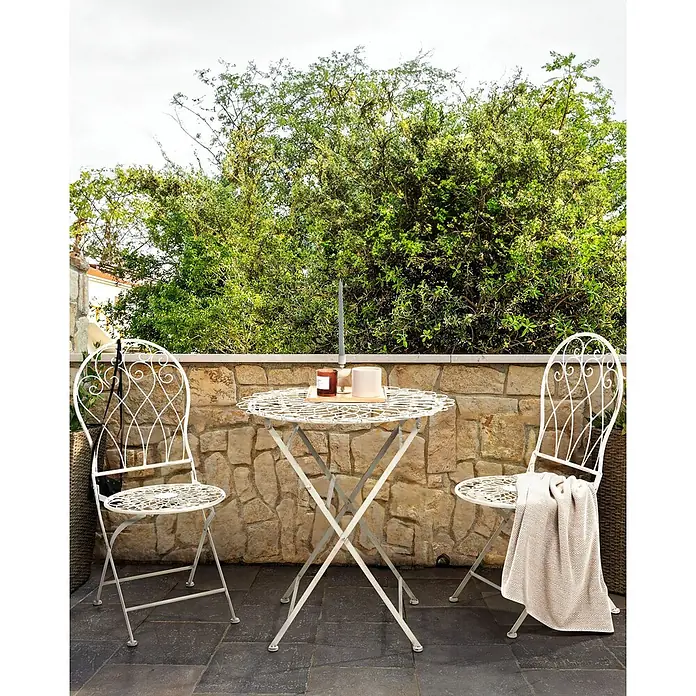 2er-Set Gartenstuhl STIFFE in Weiß -  - Weißes Metall-Bistro-Set für den Außenbereich mit verziertem Tisch und zwei Stühlen auf einer Terrasse, dekoriert mit Kerzen und Decke, an einer Steinwand.