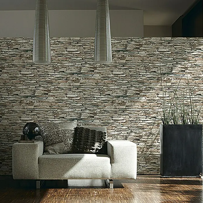 A.S. Création Vliestapete SteinNatur, Steinoptik, 10,05 x 0,53 m - Wohnzimmer mit Steinwand, grauem Sessel mit Kissen und einer dunklen Pflanzbox mit hohen Gräsern. Elegantes und modernes Design.