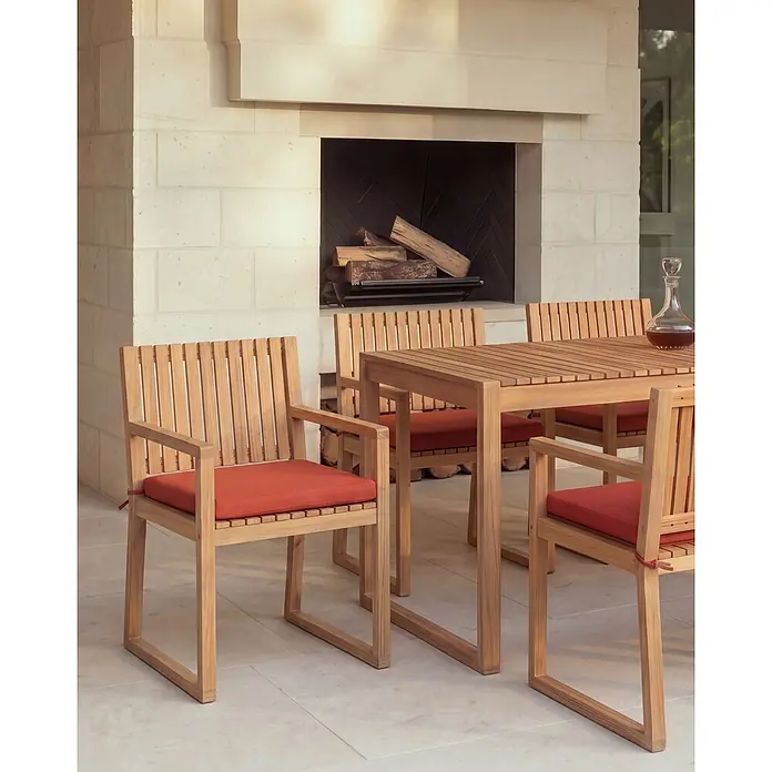 Beliani Gartenstuhl-Set SASSARI PREMIUM - 4 Stk., zertifiziertes Akazienholz, Braun, B x T x H: 54 x 59 x 80 cm - Teak-Outdoor-Essgruppe mit Lattenrosttisch und Stühlen, leuchtend orangefarbenen Kissen, auf einer Terrasse vor einem Steinkamin.