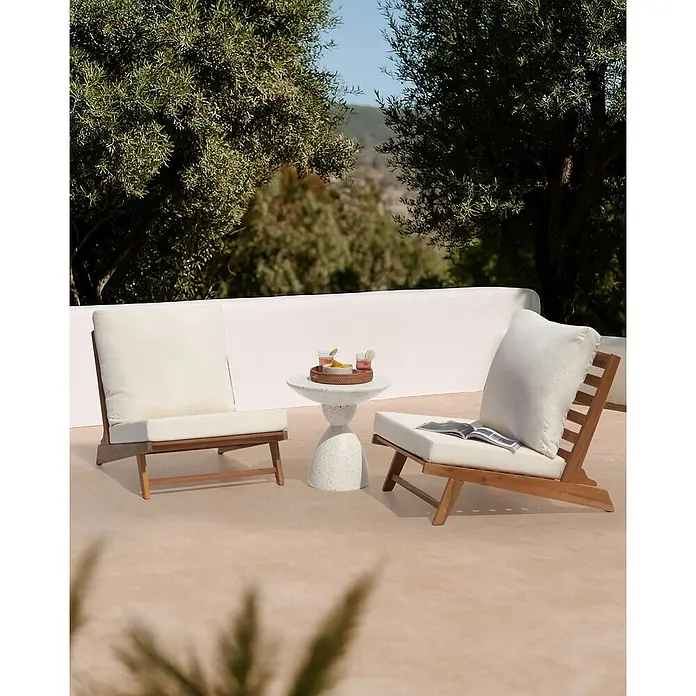 Beliani Gartenstuhl-Set MICCIANO - 2 Stk., Akazienholz, Braun, B x T x H: 70 x 91 x 71 cm - Zwei Holzliegestühle mit weißen Kissen und ein runder, heller Beistelltisch mit Getränken auf einer Terrasse.