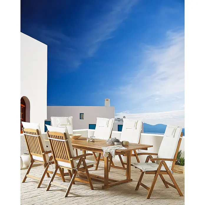 12er-Set Gartenstuhl JAVA in Braun/Weiß -  - Hölzerne Outdoor-Essgruppe mit weißen Kissen auf einer Terrasse, mit Blick auf weiße Gebäude und das Meer.