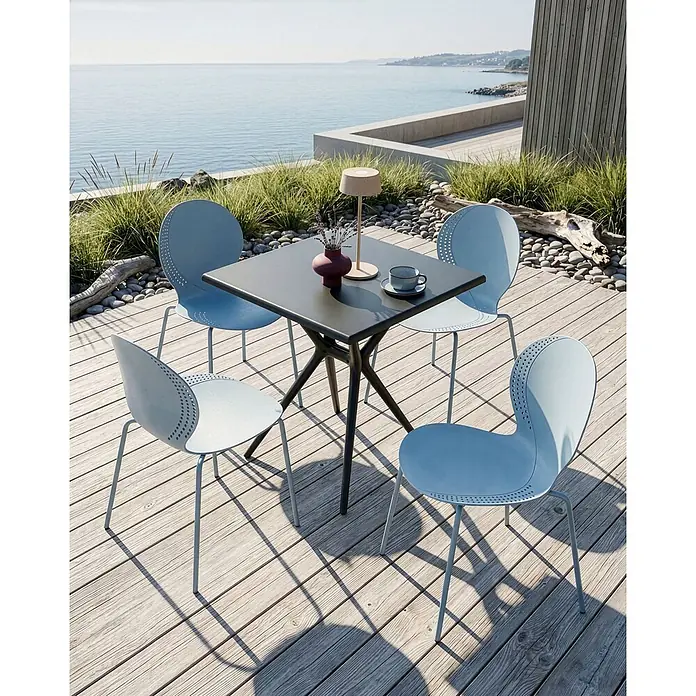 4er-Set Esszimmerstuhl MILOS in Blau -  - Ein dunkler quadratischer Tisch und vier hellblaue Stühle mit perforierten Rückenlehnen auf einer Holzterrasse mit Meerblick.