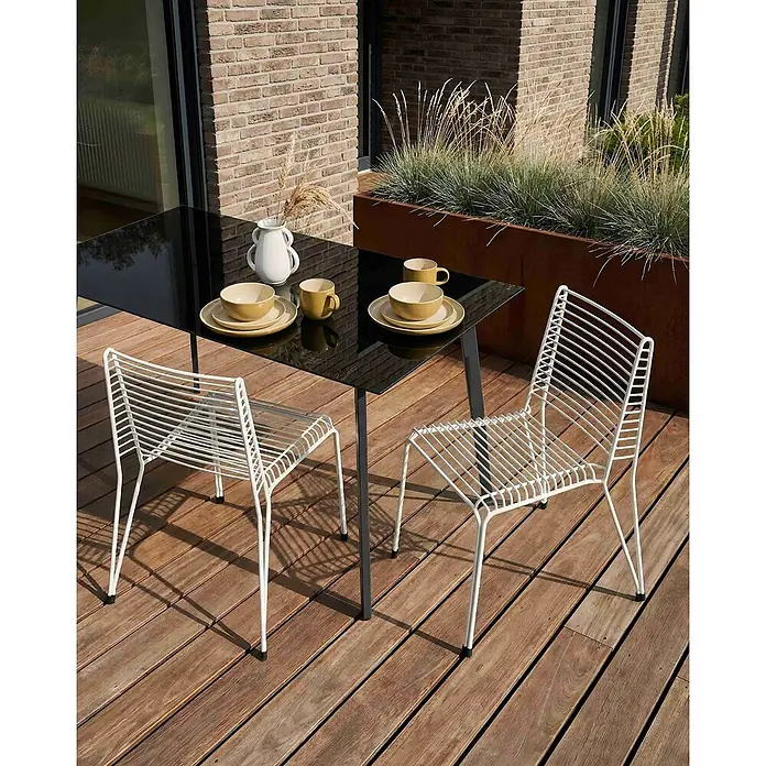 2er-Set Gartenstuhl CHIEVE in Weiß -  - Essgarnitur für draußen auf Holzterrasse: schwarzer Tisch mit gelbem Geschirr und zwei weißen Stühlen aus Metallstreben.