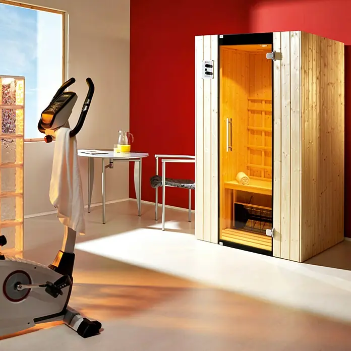Weka Infrarotkabine Tanilla 1 - 99 x 108 x 190 cm, 1.700 W, 4 Keramikstrahler - Einrichtung mit einer Sauna, einem Heimtrainer, einem Tisch und einem Stuhl. Die Sauna ist aus hellem Holz gefertigt und hat eine Glastür.