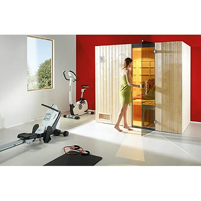 Weka Infrarot-Sauna-Kombikabine Uppsala - Therm-Flächenstrahler + 3,6 kW Saunaofen, 121 x 212 x 199 cm - Eine Frau betritt eine Sauna in einem Raum mit Fitnessgeräten. Die Sauna ist aus hellem Holz gefertigt und hat eine Glastür.