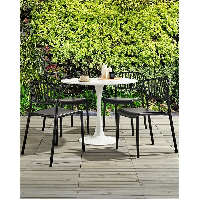 Beliani Gartenstuhl-Set GELA - 4 Stk., Polypropylen, Stapelbar, Schwarz, B x T x H: 45 x 50 x 78 cm - Ein Outdoor-Essset mit einem weißen Rundtisch und vier schwarzen Stühlen auf einer Holzterrasse, umgeben von grünen Pflanzen.