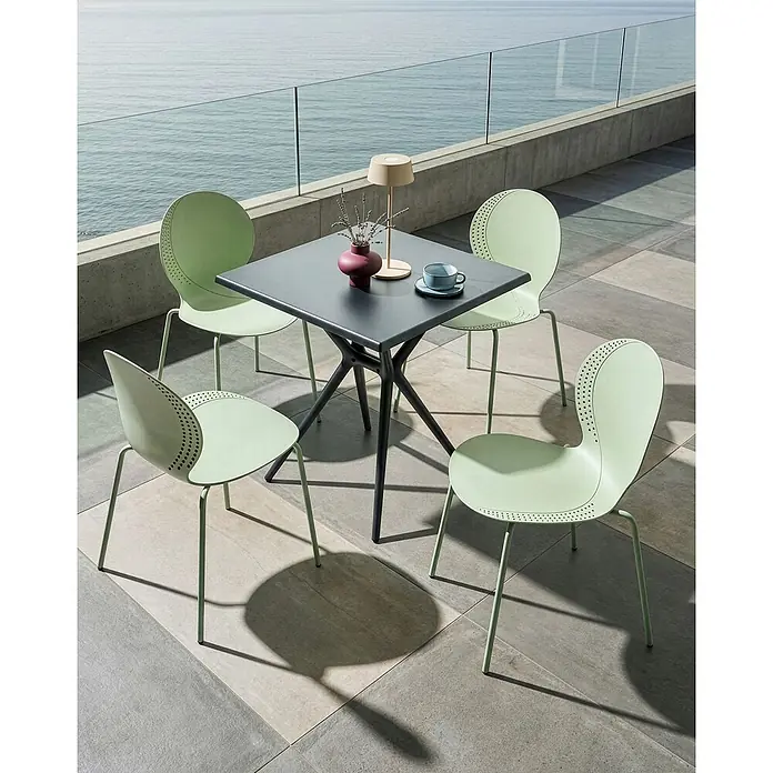 4er-Set Esszimmerstuhl MILOS in Grün -  - Quadratischer dunkelgrauer Gartentisch mit einzigartigem Fuß und vier hellgrünen perforierten Stühlen auf einer Terrasse am Meer.