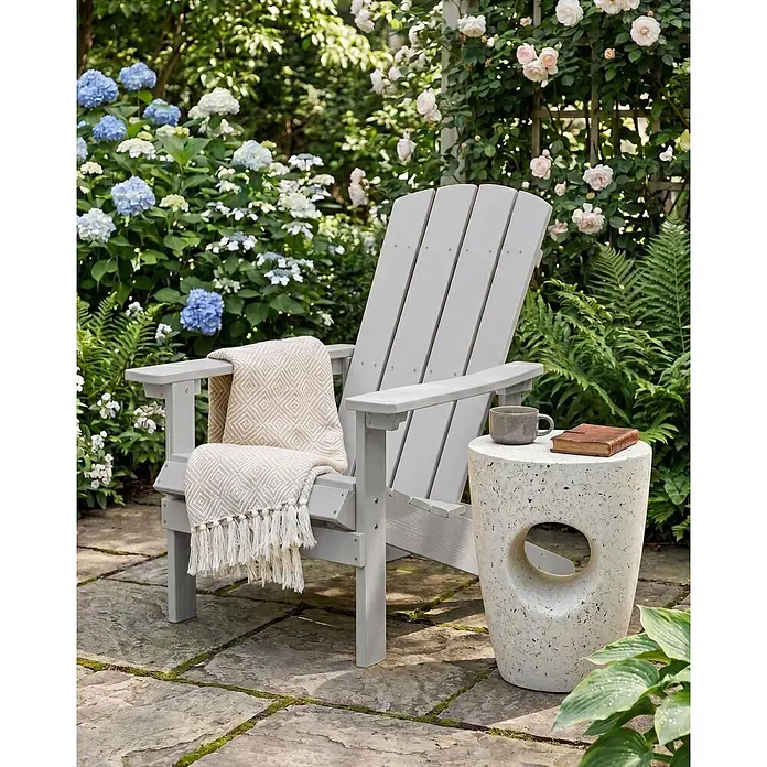 Gartenstuhl ADIRONDACK in Grau - (W) 75 x (H) 88 x - L - Hellgrauer Adirondack-Stuhl mit beigefarbener Decke auf einer Steinterrasse neben einem cremefarbenen Beistelltisch.