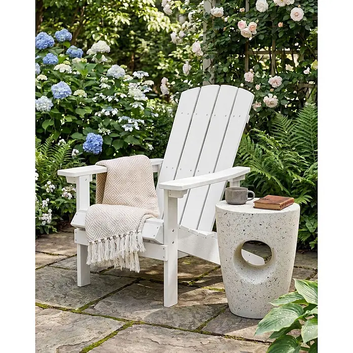 Gartenstuhl ADIRONDACK in Weiß - (W) 75 x (H) 88 x - L - Weißer Adirondack-Stuhl mit beigefarbenem Überwurf, daneben ein Beistelltisch in Betonoptik mit Tasse und Buch, in einem Garten.