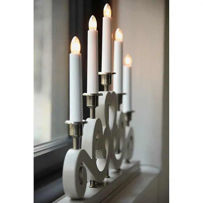 Markslöjd LED-Lichterbogen Eneby - L x B x H: 47 x 5 x 36 cm, Warmweiß, Greige - Weißer elektrischer Kerzenständer mit fünf brennenden Kerzen und Messinghaltern auf einer Fensterbank.
