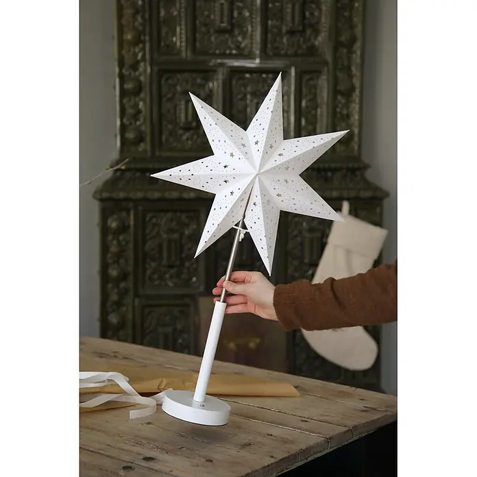 Markslöjd Standstern Noella - Ø x H: 35 x 62 cm, Weiß, LED, Akkubetrieben - Eine Person hält eine weiße Papiersternlampe mit ausgeschnittenen Sternen auf einem Ständer, bereit für festliche Dekoration.
