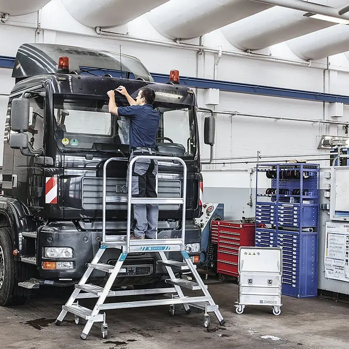 Zarges Podestleiter fahrbar mit 2x5 Leichtmetall-Stufen -  - Mobile Arbeitsplattform (Zarges) aus Aluminium mit Rollen, die einen Arbeiter am LKW unterstützt. Eine Aluminium-Transportbox ist ebenfalls sichtbar.