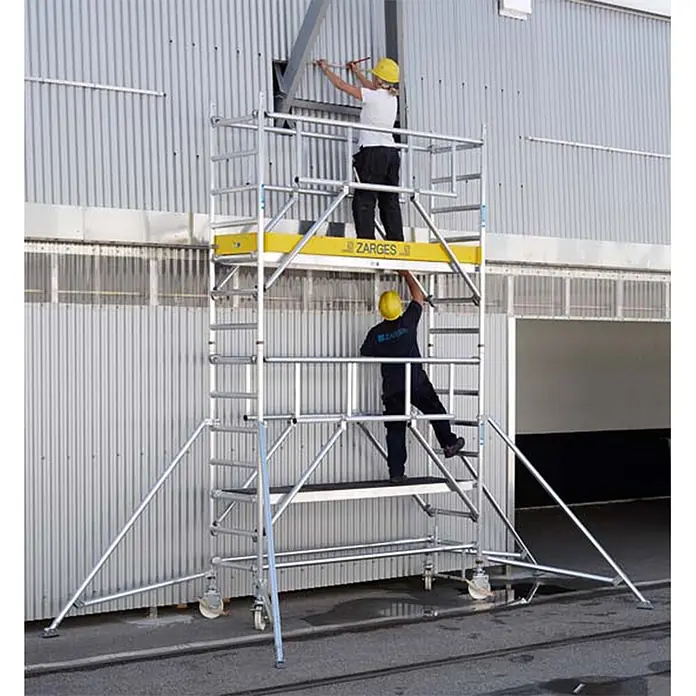 Zarges Fahrgerüst MultiTower S-PLUS 1T mit Auslegern 2,50m Plattformlänge 11,20m AH -  - Aluminium-Fahrgerüst mit gelben Plattformen, von zwei Arbeitern zur Fassadenarbeit an einem Gebäude genutzt.
