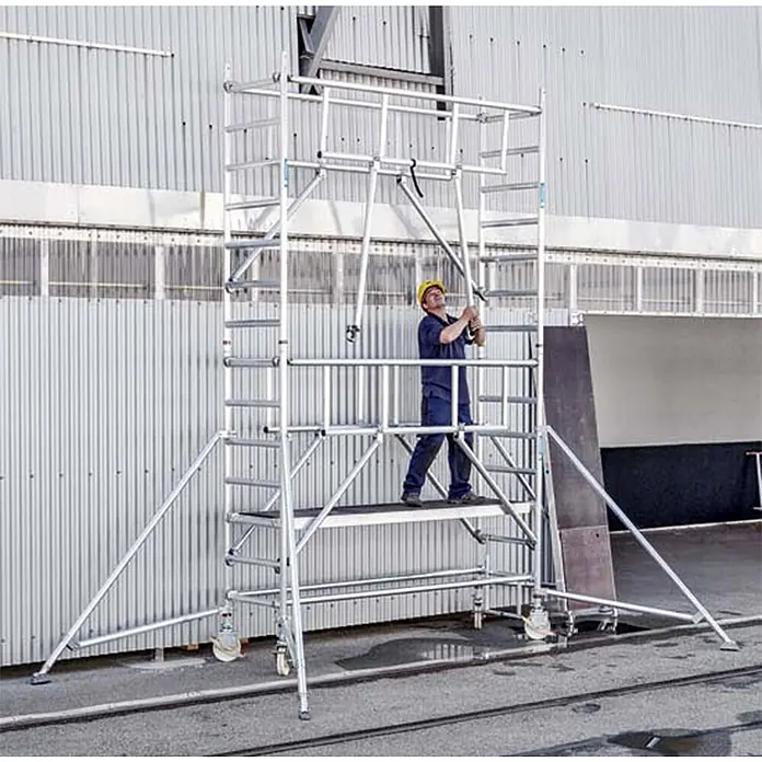 Zarges Fahrgerüst MultiTower S-PLUS 1T mit Auslegern 2,50m Plattformlänge 7,30m AH -  - Arbeiter auf einem silbernen Aluminium-Rollgerüst mit Auslegern und Rollen für Arbeiten in der Höhe, neben einem Gebäude.